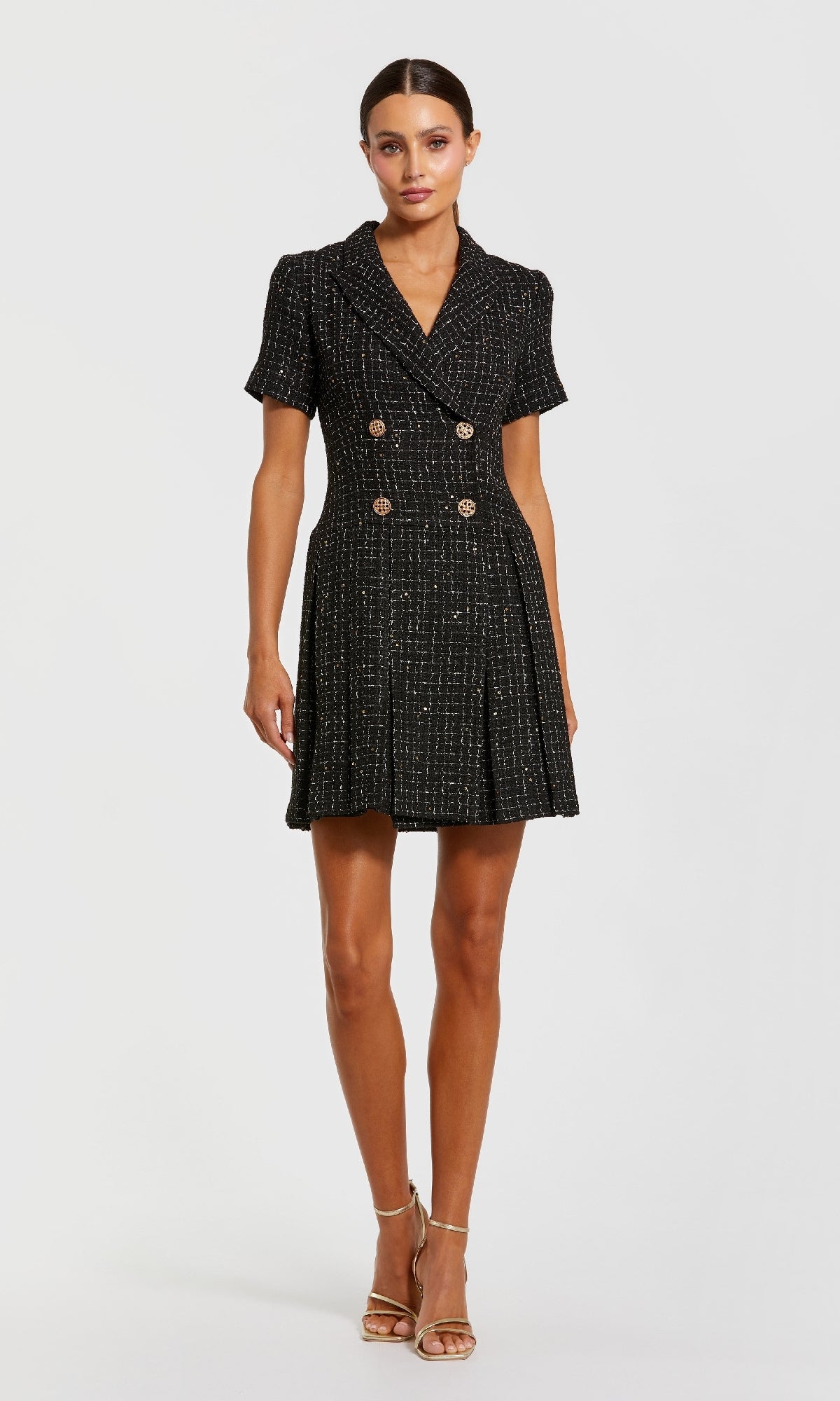 A woman in strappy heels wears the Mac Duggal 81200 Tweed-Blazer Short Party Dress—black tweed with gold buttons and pleated skirt—standing against a plain light background.